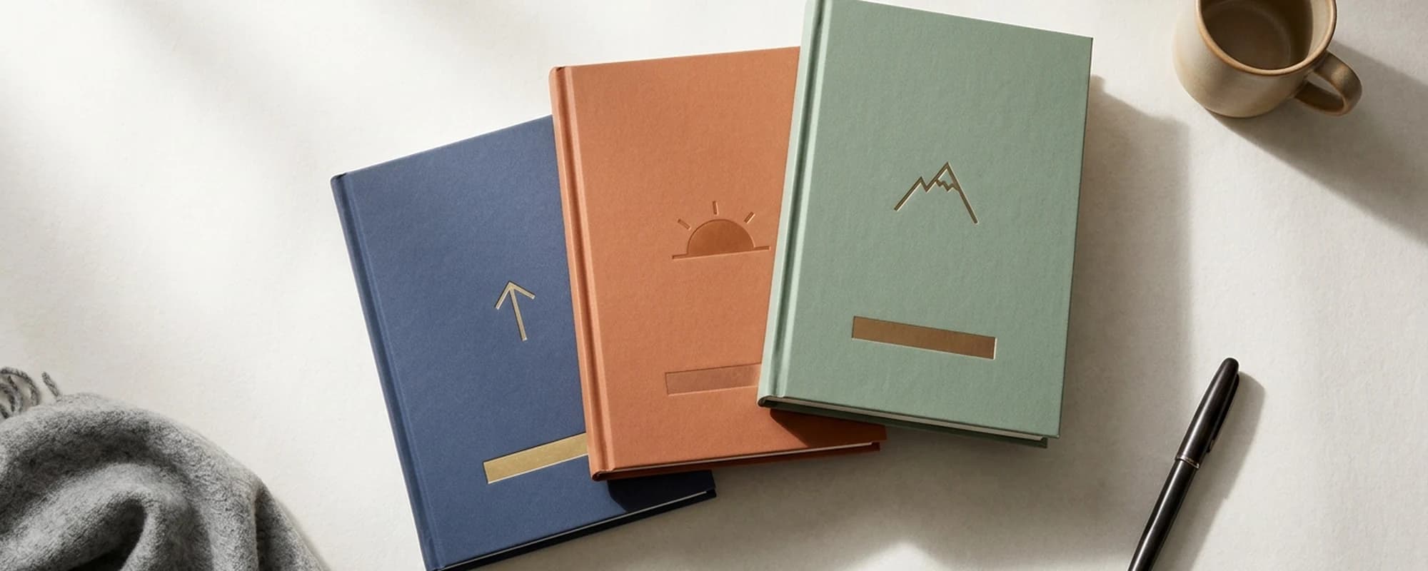 Three hardcover self-help books fanned on a soft cream surface. Each has one solid color cover with a single iconic visual: arrow, sunrise, mountain