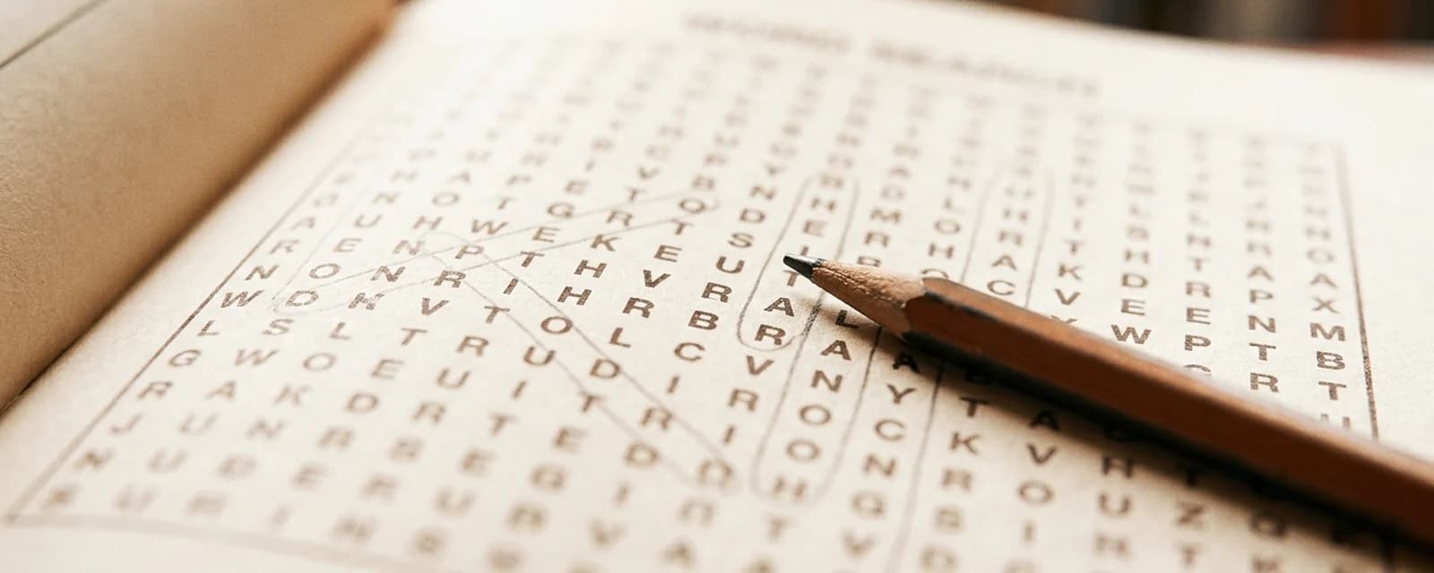 Close-up of an open word search book showing a partially solved grid with a pencil and faint diagonal lines marking found words